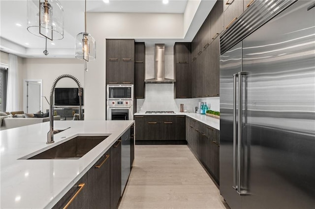 kitchen with sink, built in appliances, light wood-type flooring, pendant lighting, and wall chimney range hood