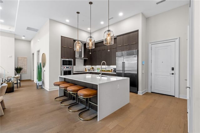 kitchen with a breakfast bar area, built in appliances, dark brown cabinets, a kitchen island with sink, and backsplash