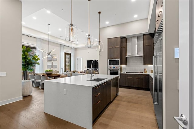 kitchen with sink, a chandelier, a center island with sink, appliances with stainless steel finishes, and wall chimney range hood