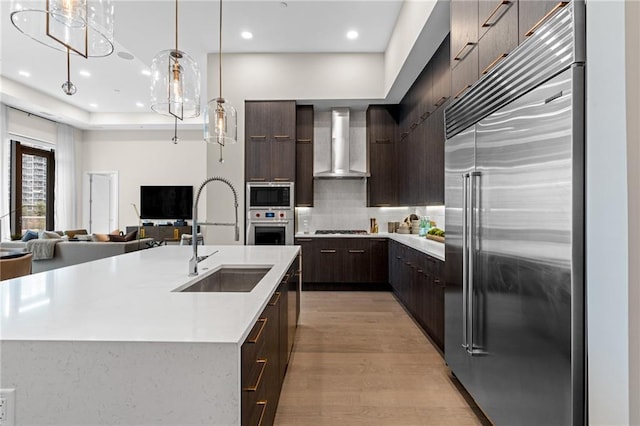 kitchen with built in appliances, sink, wall chimney exhaust hood, and a center island with sink