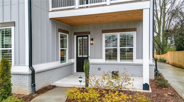 view of exterior entry with brick siding, board and batten siding, a balcony, and fence