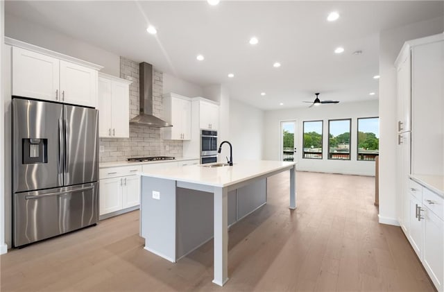 kitchen featuring white cabinets, an island with sink, wall chimney range hood, stainless steel appliances, and light hardwood / wood-style floors