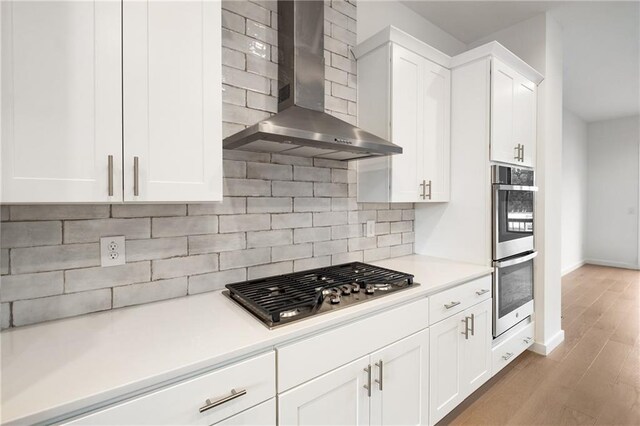 kitchen featuring light wood-type flooring, tasteful backsplash, wall chimney range hood, white cabinetry, and appliances with stainless steel finishes