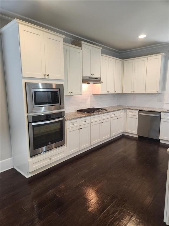 2439 Muirfield Pl, Atlanta, GA 30337 - kitchen featuring appliances with stainless steel finishes, under cabinet range hood, light stone countertops, dark wood finished floors, and decorative backsplash