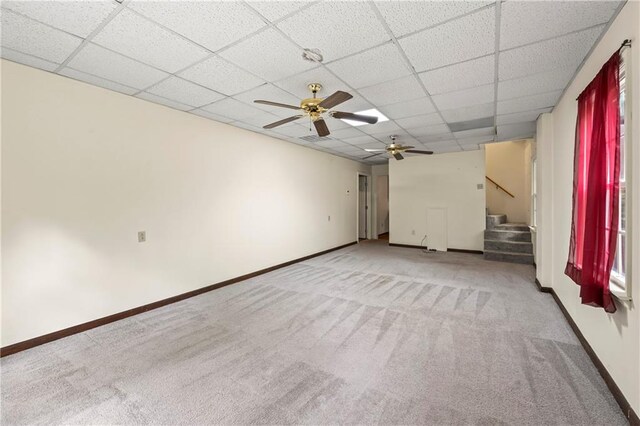 carpeted empty room with a drop ceiling, stairs, ceiling fan, and baseboards