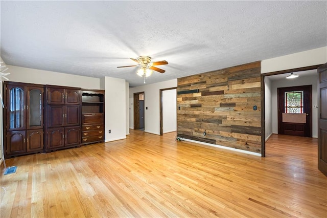 unfurnished living room with ceiling fan, light wood-style flooring, baseboards, a textured ceiling, and wooden walls