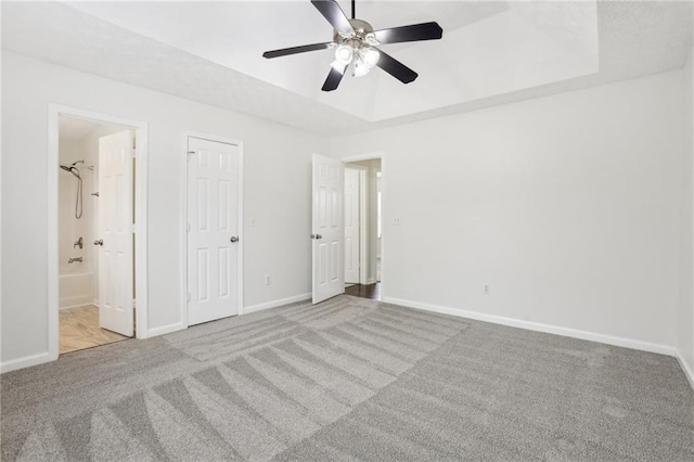 unfurnished bedroom with ceiling fan, light colored carpet, a tray ceiling, and ensuite bath