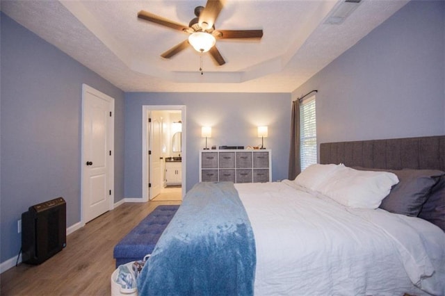 bedroom featuring ceiling fan, ensuite bath, a tray ceiling, and light hardwood / wood-style flooring