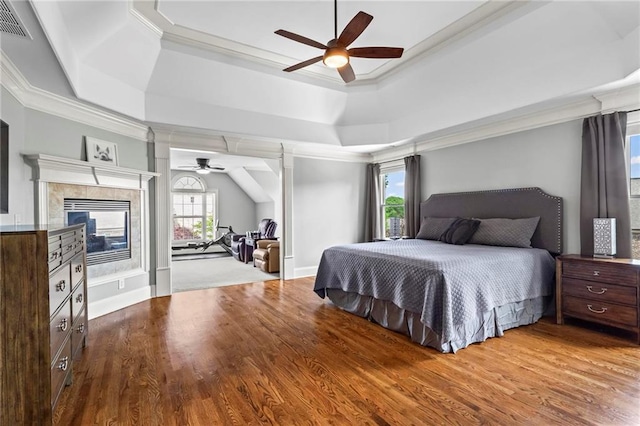 bedroom featuring ornamental molding, wood finished floors, a raised ceiling, a tiled fireplace, and ceiling fan