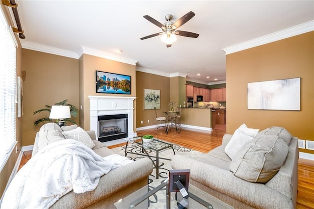 living room with ornamental molding, light wood-style flooring, baseboards, ceiling fan, and a fireplace with flush hearth