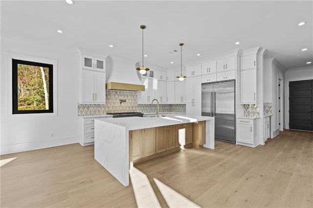 kitchen with premium range hood, a large island with sink, built in refrigerator, decorative backsplash, and white cabinetry