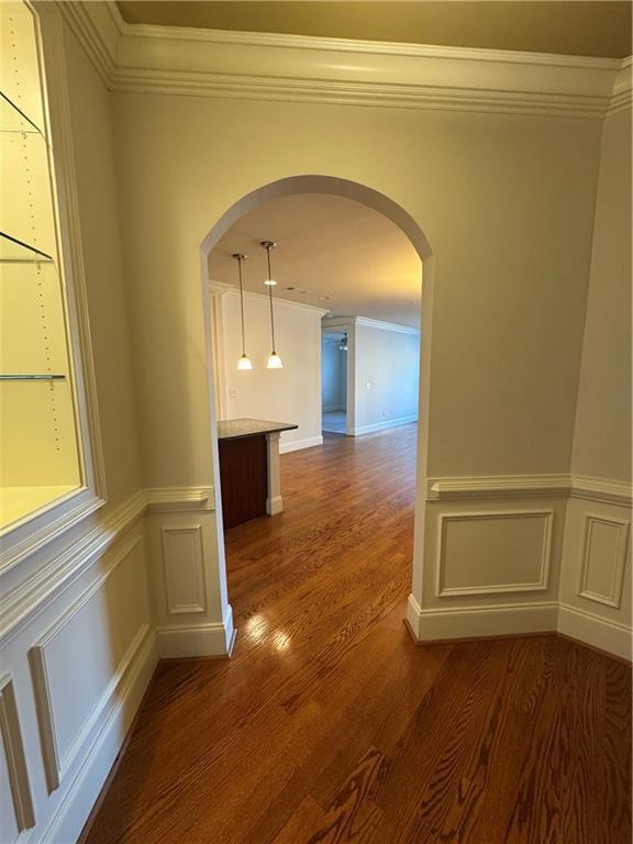hallway featuring arched walkways, wainscoting, dark wood-style flooring, crown molding, and a decorative wall