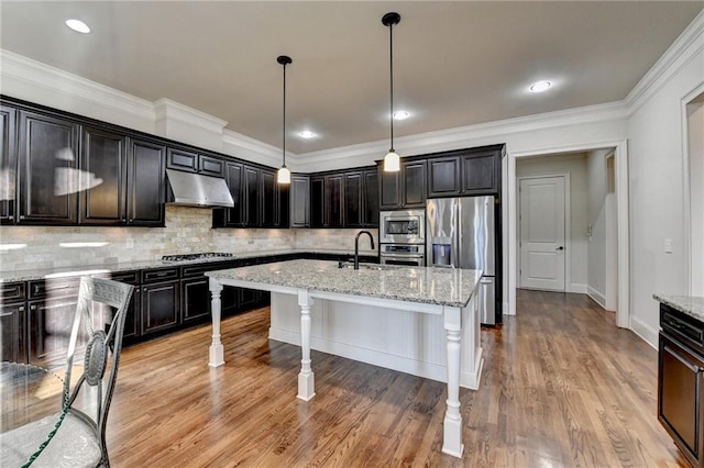 kitchen featuring decorative backsplash, appliances with stainless steel finishes, an island with sink, under cabinet range hood, and a kitchen bar