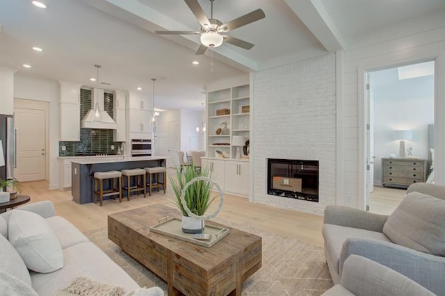 living area featuring light wood finished floors, a brick fireplace, ceiling fan, beamed ceiling, and recessed lighting