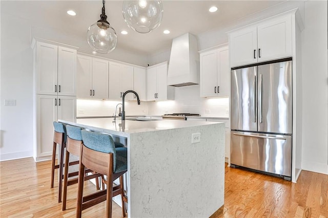 kitchen with sink, stainless steel fridge, premium range hood, white cabinetry, and a center island with sink