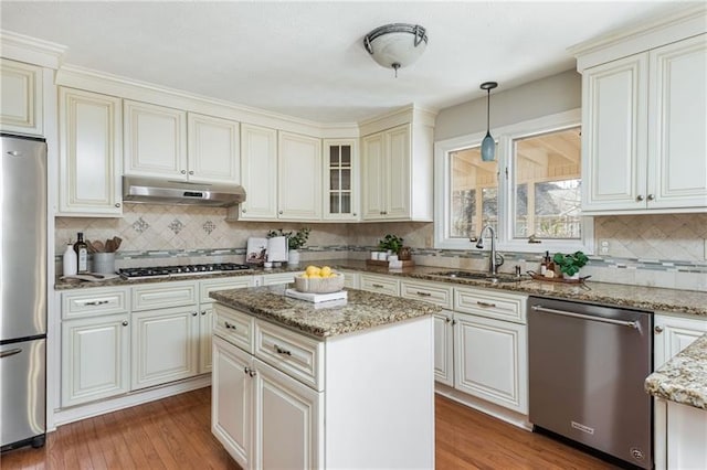 kitchen with under cabinet range hood, light stone counters, wood finished floors, stainless steel appliances, and a sink