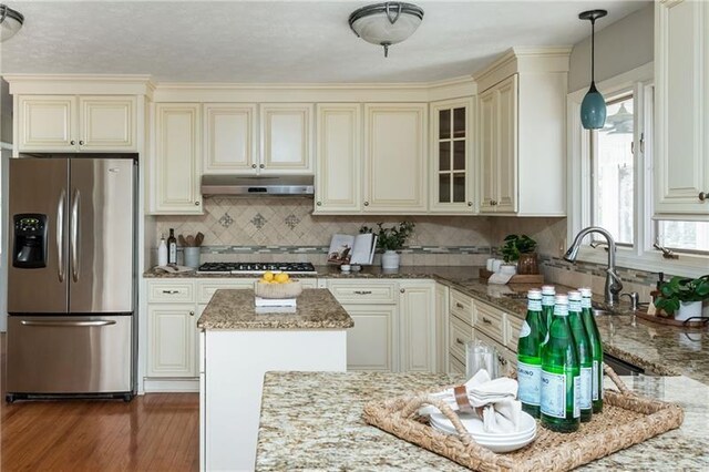 kitchen with dark wood-type flooring, cream cabinetry, under cabinet range hood, light stone counters, and stainless steel appliances