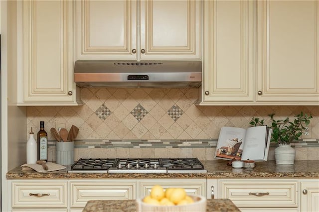 kitchen featuring under cabinet range hood, cream cabinetry, dark stone countertops, and stainless steel gas cooktop