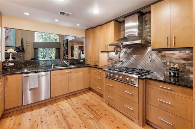 kitchen featuring dark stone countertops, light hardwood / wood-style flooring, wall chimney range hood, and appliances with stainless steel finishes