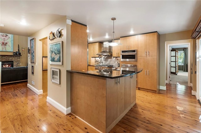 kitchen with wall chimney exhaust hood, hanging light fixtures, a stone fireplace, dark stone counters, and light hardwood / wood-style floors