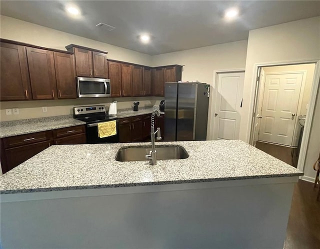 kitchen featuring light stone counters, dark brown cabinetry, stainless steel appliances, a sink, and a center island with sink