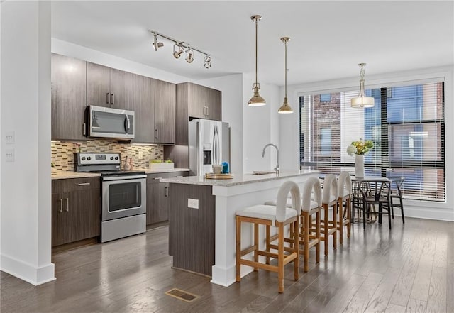 kitchen with appliances with stainless steel finishes, dark brown cabinets, dark hardwood / wood-style floors, and a kitchen island with sink