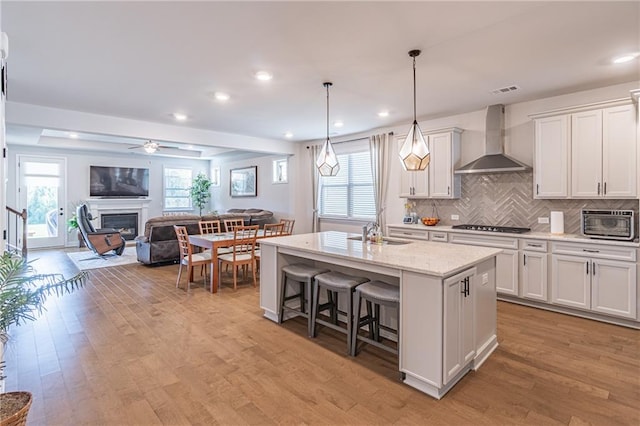 kitchen with wall chimney exhaust hood, decorative light fixtures, a kitchen island with sink, and white cabinetry