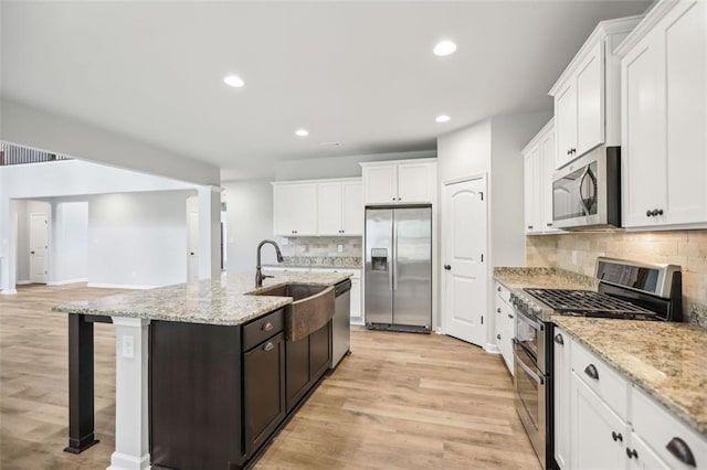 kitchen with backsplash, a kitchen island with sink, light stone countertops, appliances with stainless steel finishes, and light hardwood / wood-style floors