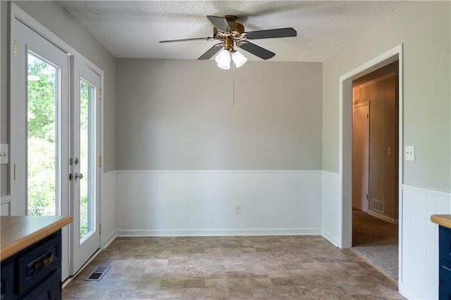 unfurnished room with healthy amount of natural light, a ceiling fan, a textured ceiling, a wainscoted wall, and stone finish floors
