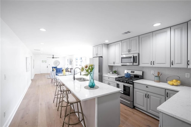 kitchen featuring ceiling fan, a center island with sink, appliances with stainless steel finishes, gray cabinetry, and a breakfast bar area