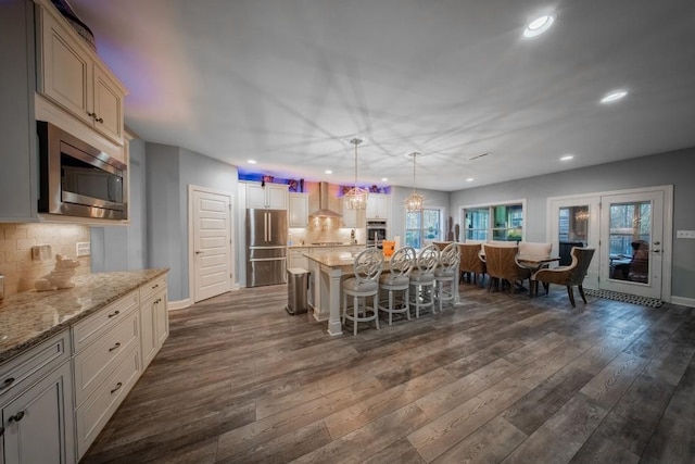 kitchen with wall chimney exhaust hood, appliances with stainless steel finishes, a kitchen breakfast bar, a kitchen island, and pendant lighting