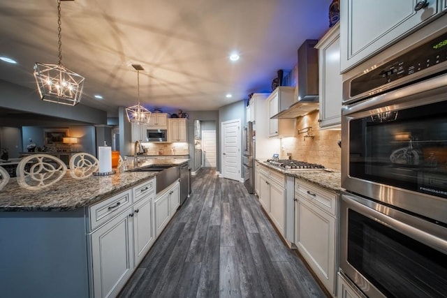 kitchen with appliances with stainless steel finishes, pendant lighting, an island with sink, sink, and wall chimney exhaust hood