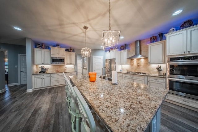 kitchen featuring appliances with stainless steel finishes, pendant lighting, a large island, light stone countertops, and wall chimney range hood