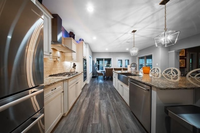 kitchen featuring wall chimney exhaust hood, light stone counters, a large island with sink, pendant lighting, and stainless steel appliances