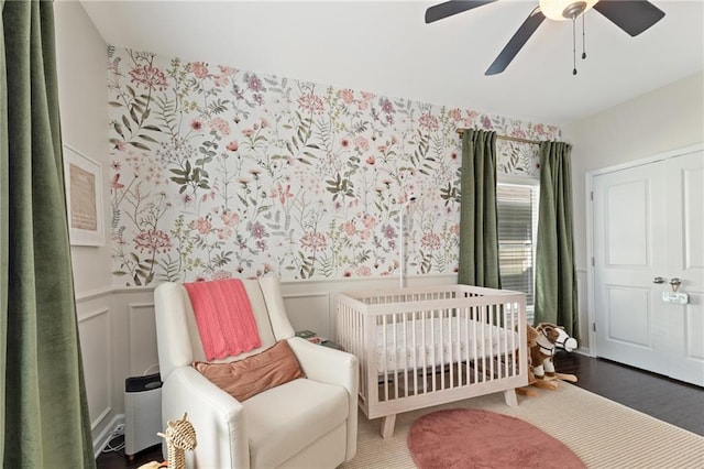 bedroom with a wainscoted wall, a crib, wallpapered walls, ceiling fan, and a decorative wall