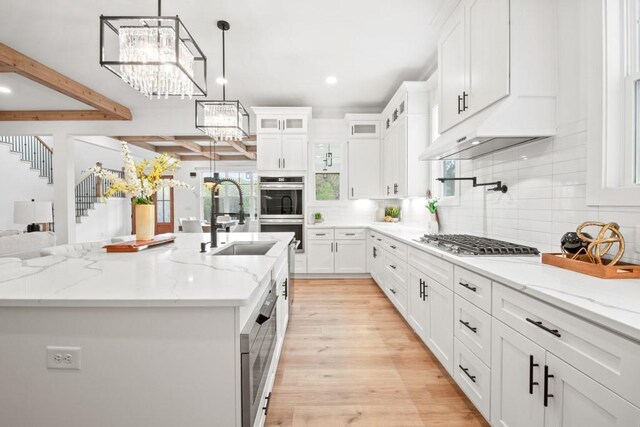 kitchen with white cabinets, appliances with stainless steel finishes, an island with sink, a sink, and an inviting chandelier
