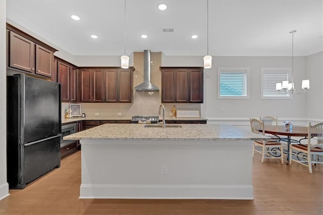 kitchen with wall chimney range hood, sink, light stone counters, an island with sink, and black fridge