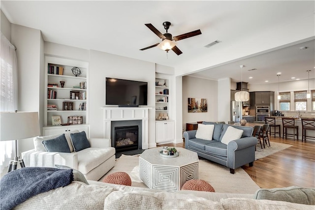 living room with built in shelves, ceiling fan, and light wood-type flooring