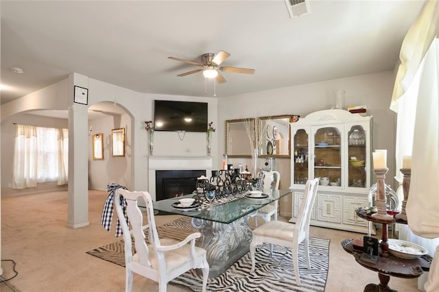 dining room with light carpet, visible vents, arched walkways, a ceiling fan, and a lit fireplace