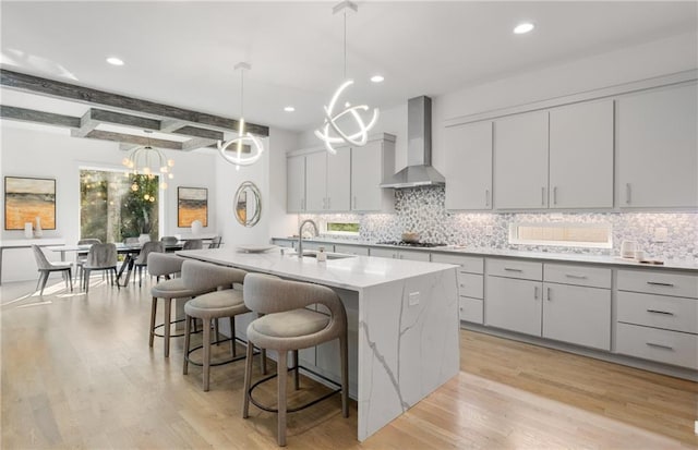 kitchen with decorative light fixtures, wall chimney range hood, beamed ceiling, sink, and a center island with sink