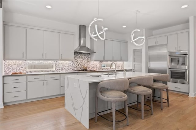 kitchen with wall chimney range hood, light wood-type flooring, pendant lighting, an island with sink, and stainless steel appliances