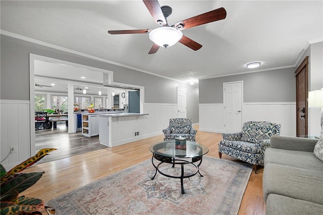 living room featuring ornamental molding, a wainscoted wall, light wood finished floors, and a ceiling fan