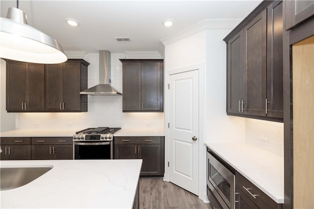 kitchen featuring decorative backsplash, dark brown cabinetry, wall chimney range hood, light wood-type flooring, and appliances with stainless steel finishes