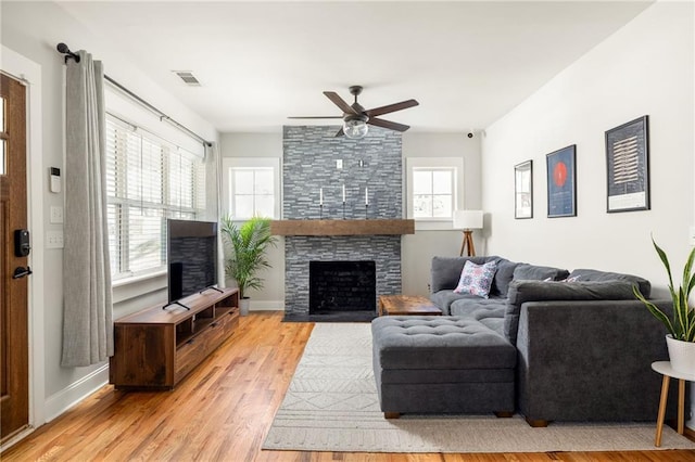 living room featuring light wood finished floors, visible vents, baseboards, ceiling fan, and a stone fireplace