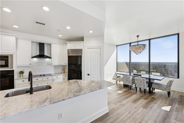 kitchen with a sink, white cabinetry, black appliances, light stone countertops, and wall chimney exhaust hood