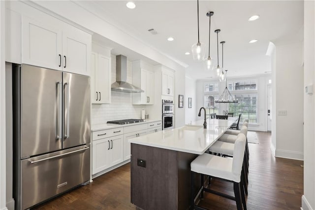 kitchen featuring appliances with stainless steel finishes, decorative light fixtures, white cabinets, a kitchen island with sink, and wall chimney range hood