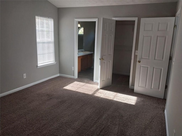 unfurnished bedroom with dark colored carpet, ensuite bathroom, a walk in closet, and vaulted ceiling