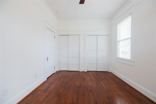 unfurnished bedroom featuring two closets, dark wood-type flooring, crown molding, baseboards, and ceiling fan