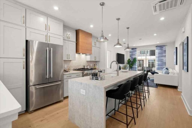 kitchen with a kitchen island with sink, hanging light fixtures, stainless steel appliances, white cabinetry, and light hardwood / wood-style floors