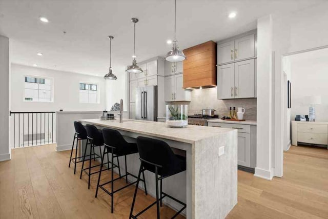 kitchen with high quality fridge, custom range hood, light wood-style floors, a kitchen island with sink, and backsplash